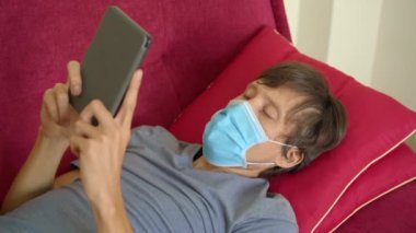 E-kitap ve online eğitim. Bir adam karantina sırasında evde elektronik kitap okuyor. Tıbbi maskeli bir adam evde tek başına kaldı.