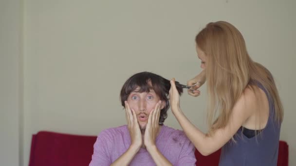 En ung kvinna klipper sin pojkvän eller make på grund av nedlåsning. Hon försöker följa videoinstruktionerna. Han är rädd att hans frisyr ska bli hemsk. Begreppet social isolering — Stockvideo