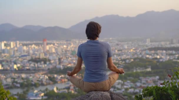 Egy fiatalember meditál egy hegyen egy nagyváros felett. A belső béke fogalma egy nagyvárosban — Stock videók