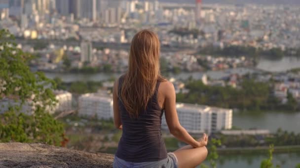 Una joven medita en una montaña sobre una gran ciudad. Concepto de paz interior en una gran ciudad — Vídeos de Stock