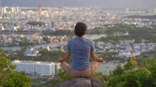 Genç bir adam büyük bir şehrin tepesindeki bir dağda meditasyon yapıyor. Büyük bir şehirde iç huzur kavramı — Stok video