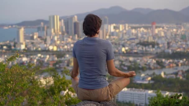 Genç bir adam büyük bir şehrin tepesindeki bir dağda meditasyon yapıyor. Büyük bir şehirde iç huzur kavramı — Stok video