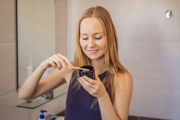young woman brushing her teeth with a black tooth paste with active charcoal, and black tooth brush in her bathroom