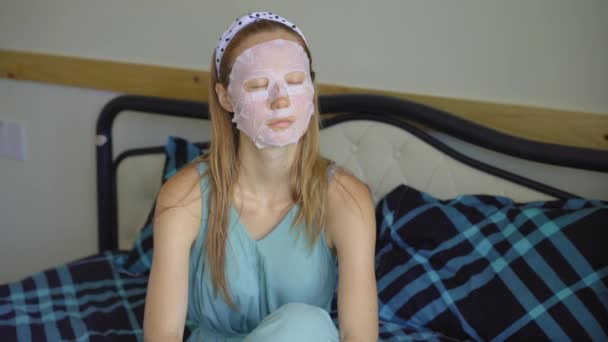 En ung kvinna sitter på en säng med en kosmetisk ansiktsmask i ansiktet. Hudvårdskoncept — Stockvideo