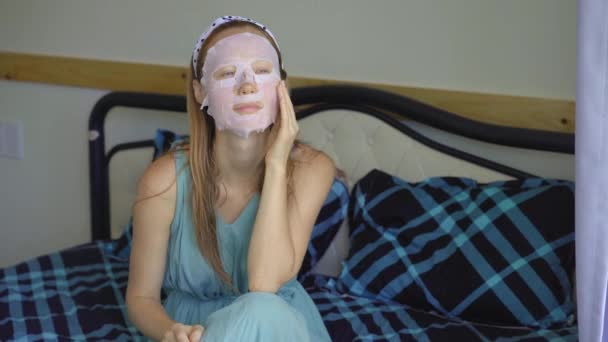 Una joven está sentada en una cama con una mascarilla cosmética en la cara. Concepto de cuidado de la piel — Vídeo de stock