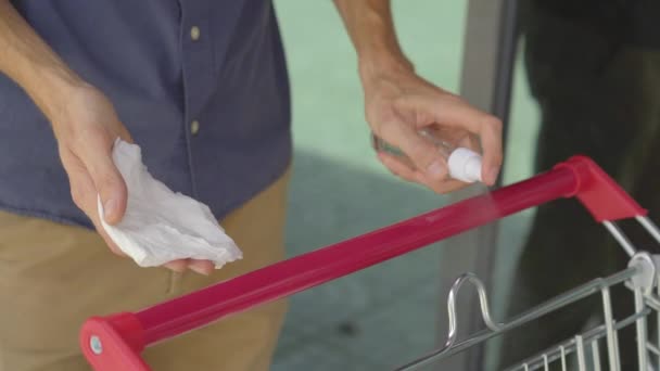 Jongeman desinfecteert een handvat van een supermarkt trolley om zichzelf te beschermen tegen contact met de infectie. Sociaal afstandelijk concept. Covid-beschermingsconcept. — Stockvideo
