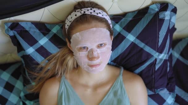 Una giovane donna sdraiata su un letto con una maschera cosmetica sul viso. Concetto di cura della pelle — Video Stock