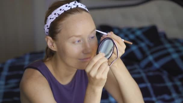 Mujer joven haciendo maquillaje mientras está sentada en una cama — Vídeos de Stock