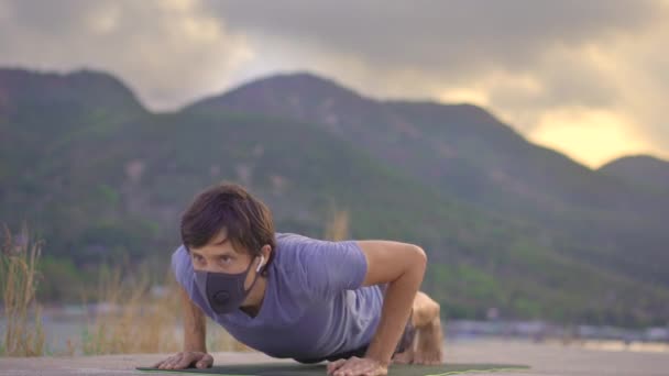 Un joven con una mascarilla protectora hace entrenamiento físico en un río o junto al mar. Actividad física durante la cuarentena. Brote de Covid-19 — Vídeos de Stock