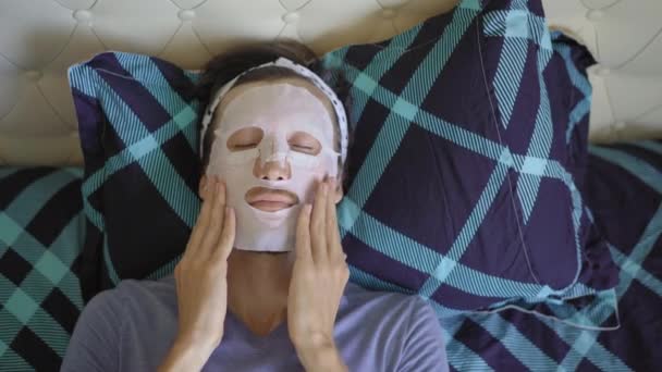 Un joven acostado en una cama con una mascarilla cosmética en la cara. Concepto de cuidado de la piel — Vídeos de Stock