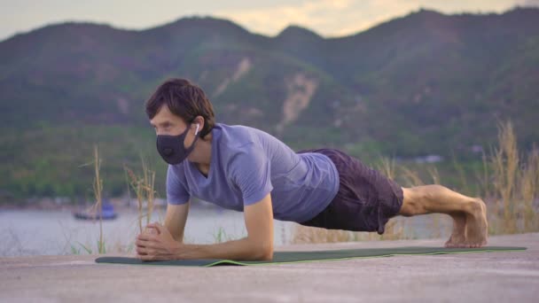 A young man wearing a protective face mask does physical training on a river or seaside. Physical activity during the quarantine. Covid-19 outbreak — Stock Video