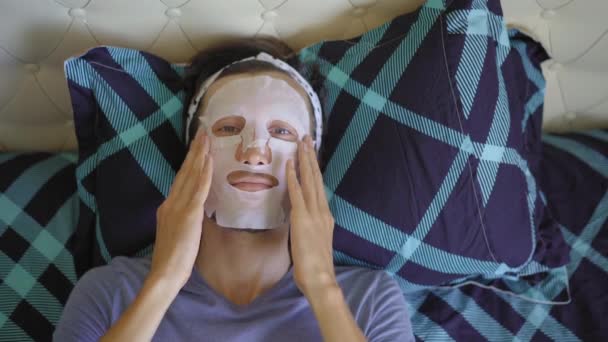 En ung man som ligger på en säng med en kosmetisk ansiktsmask i ansiktet. Hudvårdskoncept — Stockvideo