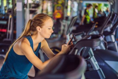 Coronavirus bitti. Karantina zayıfladı. Maskeni çıkar. Artık halka açık yerlere gidebilirsin. Spor salonunda eğitim görürken telefonu kullanan genç bir kadın. Egzersiz makinesinde oturan kadın cep telefonunu tutuyor