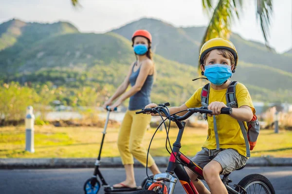 Active school kid boy and his mom in medical mask and safety helmet riding a bike with backpack on sunny day. Happy child biking on way to school. You need to go to school in a mask because of the
