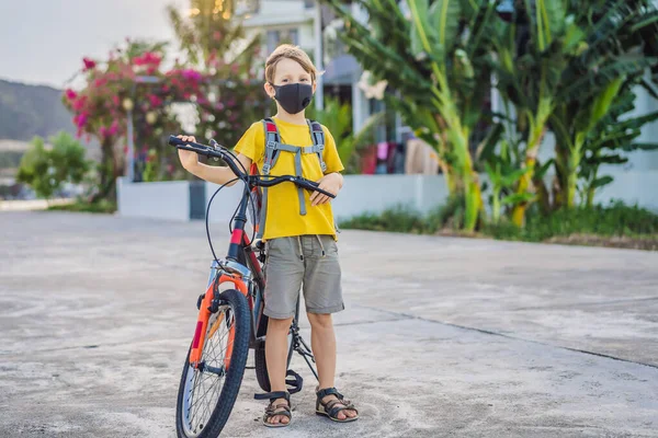 Actieve schooljongen met medisch masker op een fiets met rugzak op zonnige dag. Gelukkig kind fietsen op weg naar school. Je moet met een masker naar school vanwege de coronavirus epidemie. — Stockfoto