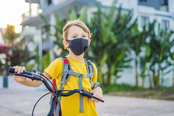Menino de escola ativa em máscara médica andando de bicicleta com mochila no dia ensolarado. Criança feliz de bicicleta a caminho da escola. Tens de ir para a escola mascarada por causa da epidemia de coronavírus. — Fotografia de Stock