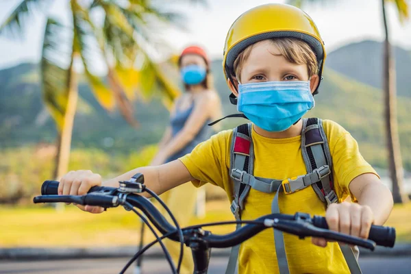 Enfant de l'école active garçon et sa mère en masque médical et casque de sécurité en vélo avec sac à dos sur la journée ensoleillée. Joyeux enfant faisant du vélo sur le chemin de l'école. Tu dois aller à l'école avec un masque à cause de la — Photo