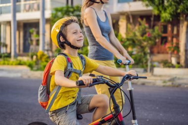 Aktif okul çocuğu ve annesi kasklı ve güneşli bir günde sırt çantasıyla bisiklet sürüyor. Okula giderken mutlu çocuk bisikleti. Çocuklar için güvenli okul yolu.