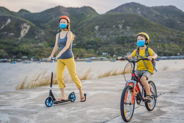 Der aktive Schuljunge und seine Mutter in medizinischer Maske und Schutzhelm fahren an sonnigen Tagen Fahrrad mit Rucksack. Glückliches Kind mit dem Fahrrad auf dem Schulweg. Sie müssen zur Schule gehen mit einer Maske wegen der — Stockfoto