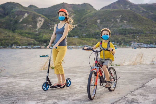 Active school kid boy and his mom in medical mask and safety helmet riding a bike with backpack on sunny day. Happy child biking on way to school. You need to go to school in a mask because of the — Stock Photo, Image