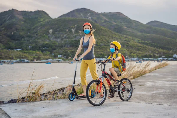 Der aktive Schuljunge und seine Mutter in medizinischer Maske und Schutzhelm fahren an sonnigen Tagen Fahrrad mit Rucksack. Glückliches Kind mit dem Fahrrad auf dem Schulweg. Sie müssen zur Schule gehen mit einer Maske wegen der — Stockfoto