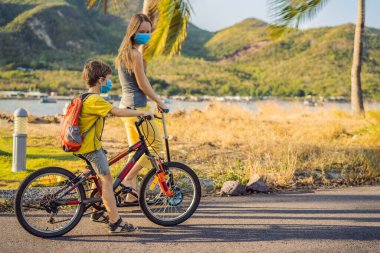 Aktif okul çocuğu ve tıbbi maskeli annesi güneşli bir günde sırt çantasıyla bisiklet sürüyorlar. Okula giderken mutlu çocuk bisikleti. Koronavirüs salgını yüzünden okula maskeyle gitmen gerekiyor.