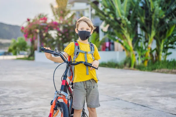 阳光明媚的日子里，戴着医疗面罩的积极的学童骑着背包骑自行车。快乐的孩子骑自行车上学的路上。3.你必须戴上口罩才能去学校，因为这种病很流行 — 图库照片