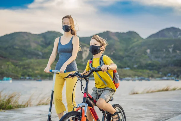 阳光灿烂的日子里，积极的学童男孩和他的妈妈戴着医疗面罩骑着一辆背着背包的自行车。快乐的孩子骑自行车上学的路上。3.你必须戴上口罩才能去学校，因为这种病很流行 — 图库照片