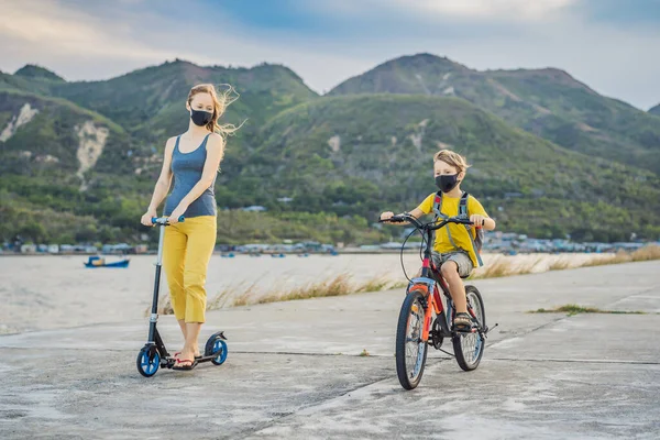 Aktív iskolás fiú és az anyja orvosi maszkban bicikliznek hátizsákkal a napsütéses napon. Boldog gyerek biciklizés úton az iskolába. Maszkban kell iskolába menned a koronavírus járvány miatt. — Stock Fotó
