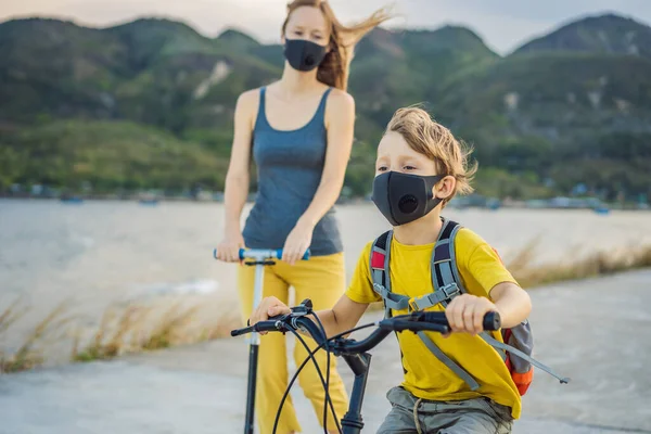 阳光灿烂的日子里，积极的学童男孩和他的妈妈戴着医疗面罩骑着一辆背着背包的自行车。快乐的孩子骑自行车上学的路上。3.你必须戴上口罩才能去学校，因为这种病很流行 — 图库照片