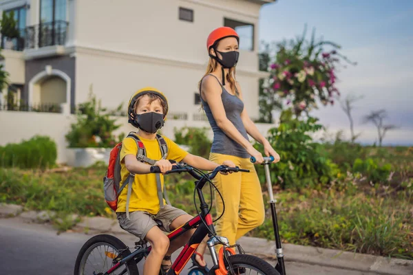 阳光明媚的日子里，积极的学童男孩和他的妈妈戴着医疗面罩，戴着安全帽骑着自行车背着背包。快乐的孩子骑自行车上学的路上。你需要带着面具去学校，因为... — 图库照片