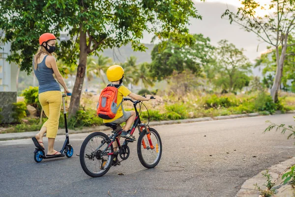 Actieve schooljongen en zijn moeder met medisch masker en veiligheidshelm op een fiets met rugzak op zonnige dag. Gelukkig kind fietsen op weg naar school. Je moet naar school gaan in een masker vanwege de — Stockfoto