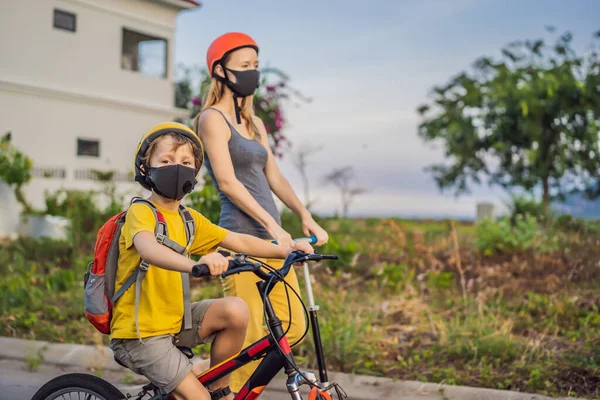 阳光明媚的日子里，积极的学童男孩和他的妈妈戴着医疗面罩，戴着安全帽骑着自行车背着背包。快乐的孩子骑自行车上学的路上。你需要带着面具去学校，因为... — 图库照片