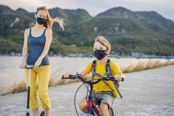 Der aktive Schuljunge und seine Mutter in medizinischer Maske auf einem Fahrrad mit Rucksack an einem sonnigen Tag. Glückliches Kind mit dem Fahrrad auf dem Schulweg. Sie müssen wegen der Coronavirus-Epidemie maskiert zur Schule gehen — Stockfoto