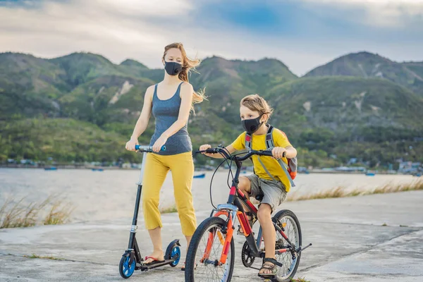 Active school kid boy and his mom in medical mask riding a bike with backpack on sunny day. Happy child biking on way to school. You need to go to school in a mask because of the coronavirus epidemic — Stock Photo, Image