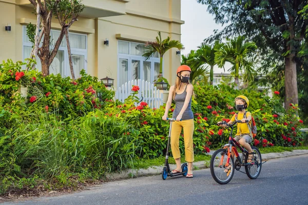 Actieve schooljongen en zijn moeder met medisch masker en veiligheidshelm op een fiets met rugzak op zonnige dag. Gelukkig kind fietsen op weg naar school. Je moet naar school gaan in een masker vanwege de — Stockfoto