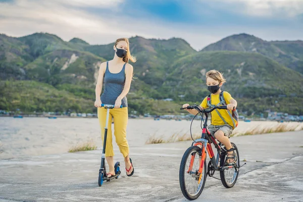 Actieve schooljongen en zijn moeder met medisch masker rijden op een fiets met rugzak op zonnige dag. Gelukkig kind fietsen op weg naar school. Je moet met een masker naar school vanwege de coronavirus epidemie. — Stockfoto