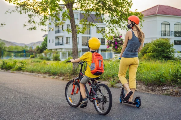 Active school kid boy and his mom in medical mask and safety helmet riding a bike with backpack on sunny day. Happy child biking on way to school. You need to go to school in a mask because of the