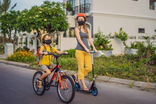 Actieve schooljongen en zijn moeder met medisch masker en veiligheidshelm op een fiets met rugzak op zonnige dag. Gelukkig kind fietsen op weg naar school. Je moet naar school gaan in een masker vanwege de — Stockfoto