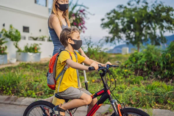 Active school kid boy and his mom in medical mask riding a bike with backpack on sunny day. Happy child biking on way to school. You need to go to school in a mask because of the coronavirus epidemic