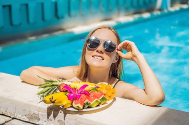 Genç bir kadın otel havuzunda dinleniyor ve meyve tabağı yiyor. Egzotik yaz diyeti. Havuz kenarında sağlıklı yiyecekler olan bacakların fotoğrafı, yukarıdan üst manzara. Tropik sahil yaşam tarzı