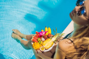 Genç bir kadın otel havuzunda dinleniyor ve meyve tabağı yiyor. Egzotik yaz diyeti. Havuz kenarında sağlıklı yiyecekler olan bacakların fotoğrafı, yukarıdan üst manzara. Tropik sahil yaşam tarzı