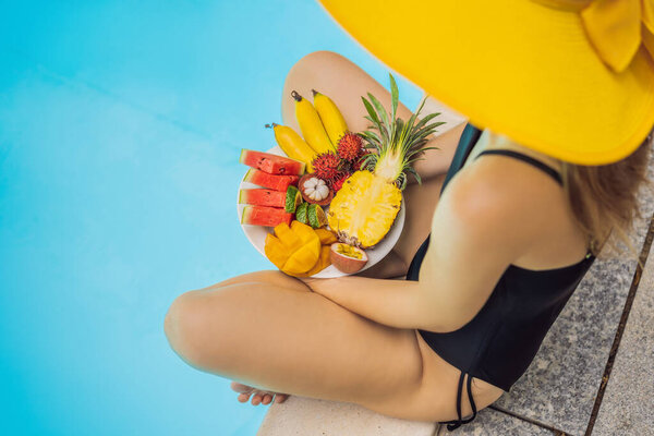 Young woman relaxing and eating fruit plate by the hotel pool. Exotic summer diet. Photo of legs with healthy food by the poolside, top view from above. Tropical beach lifestyle