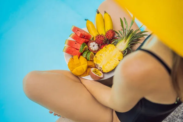 Mujer joven relajándose y comiendo plato de frutas junto a la piscina del hotel. Dieta exótica de verano. Foto de piernas con comida sana junto a la piscina, vista superior desde arriba. Estilo de vida de playa tropical — Foto de Stock