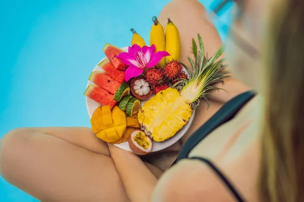 Mujer joven relajándose y comiendo plato de frutas junto a la piscina del hotel. Dieta exótica de verano. Foto de piernas con comida sana junto a la piscina, vista superior desde arriba. Estilo de vida de playa tropical — Foto de Stock