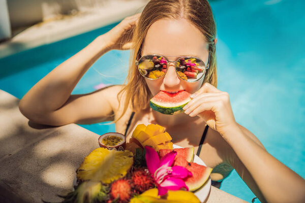 Молодая женщина отдыхает и ест фруктовую тарелку у бассейна отеля. Экзотическая летняя диета. Фото ног со здоровой пищей у бассейна, вид сверху. Тропический образ жизни

