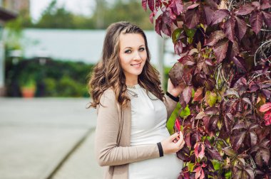 Kızıl sonbahar çitinde karnını tutan hamile bir genç kadın. Hamile kadın parkta dinleniyor..