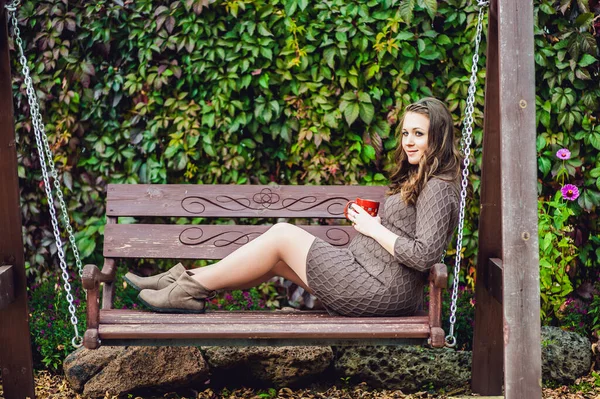 Egy terhes fiatal nő ül a hintán, egy csésze teával. terhes nő pihenés a parkban. — Stock Fotó