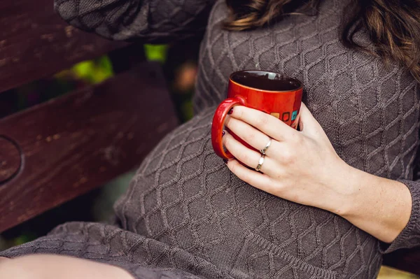 Η κοιλιά μιας εγκύου που κάθεται σε μια κούνια, με ένα φλιτζάνι τσάι. έγκυος γυναίκα χαλαρώνοντας στο πάρκο. — Φωτογραφία Αρχείου