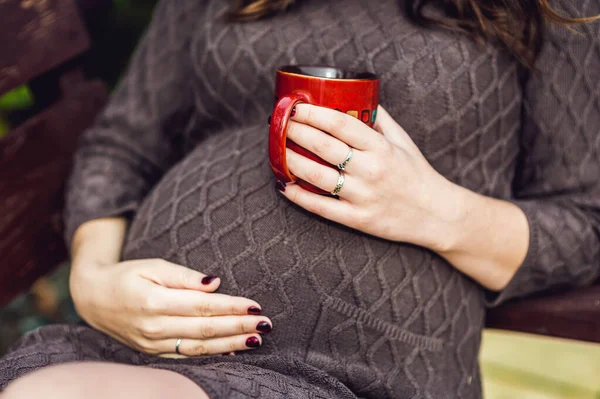 Břicho těhotné ženy sedící na houpačce s šálkem čaje. těhotná žena relaxace v parku. — Stock fotografie